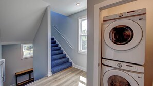 Une buanderie avec des machines à laver et des sèche-linge superposés, un escalier recouvert de moquette bleue et un banc.