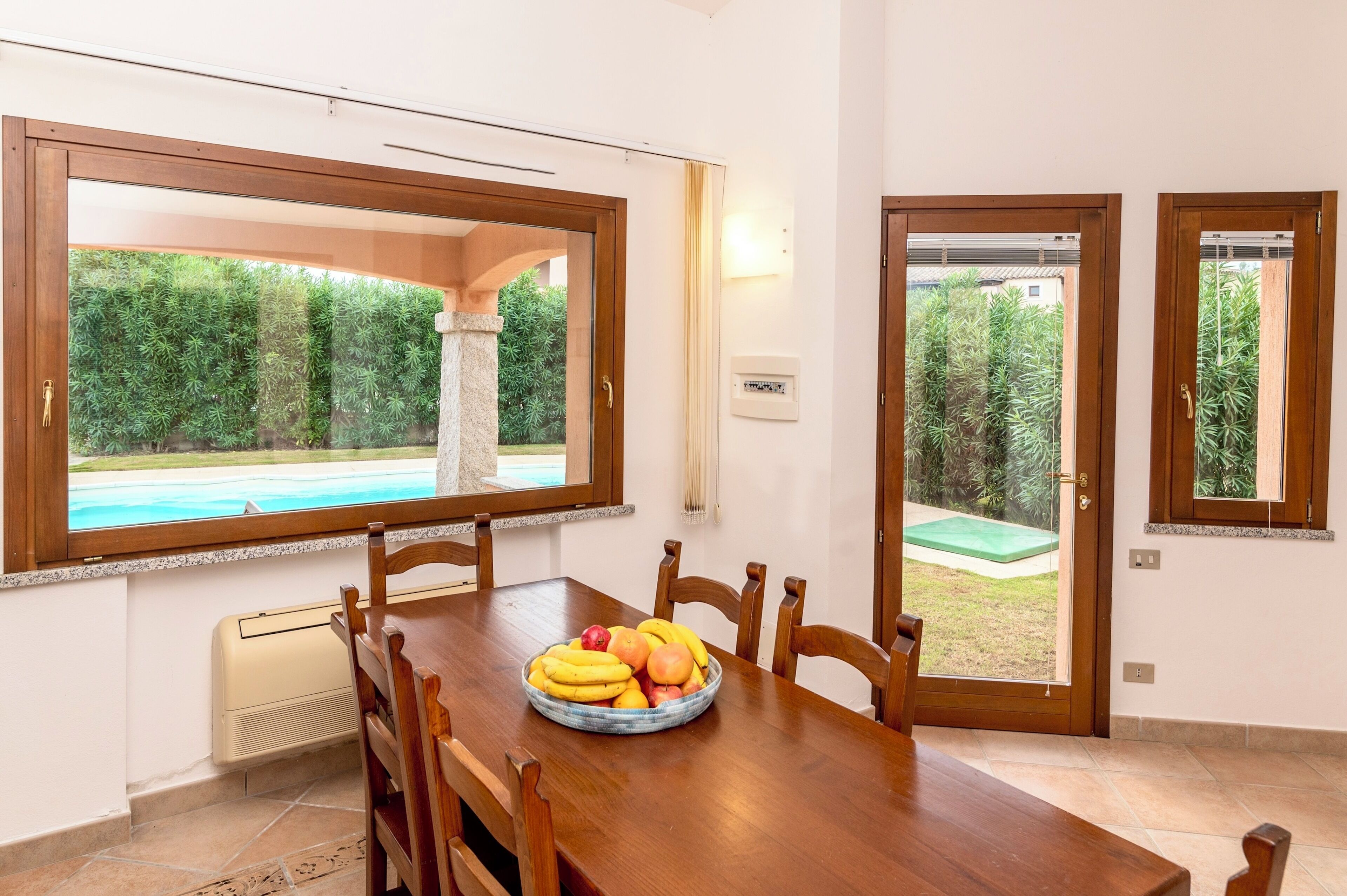 A dining area with a wooden table, chairs, and a bowl of fruit. There are windows with views of greenery and a pool.