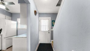 Un pasillo estrecho con una puerta blanca al final, un felpudo negro y un ventilador de techo.