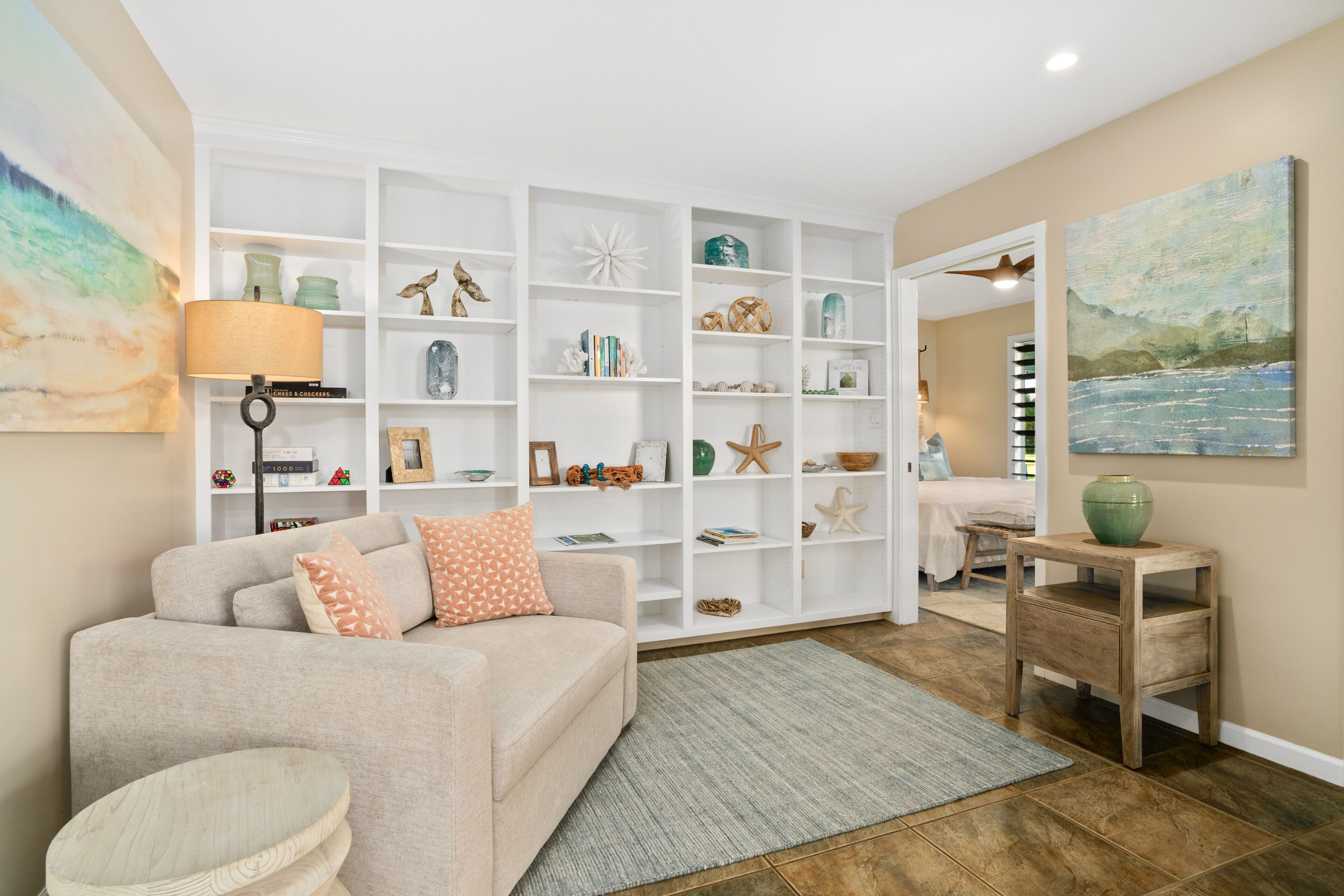 A living room with a beige sofa, a wooden side table, a bookshelf filled with decorative items, and a painting on the wall.