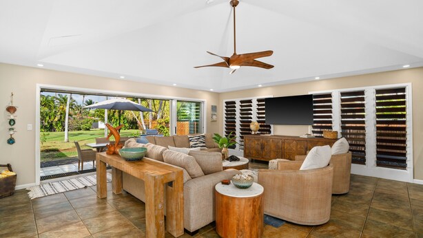 A spacious living room with a large sofa, a coffee table, and a ceiling fan. There is a large window with a view of a garden.