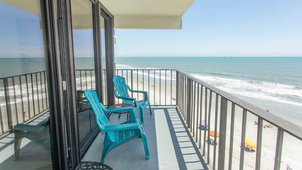 Een balkon met twee turquoise stoelen en een tafel, met uitzicht op een strand met een hek en een helder uitzicht over de oceaan.