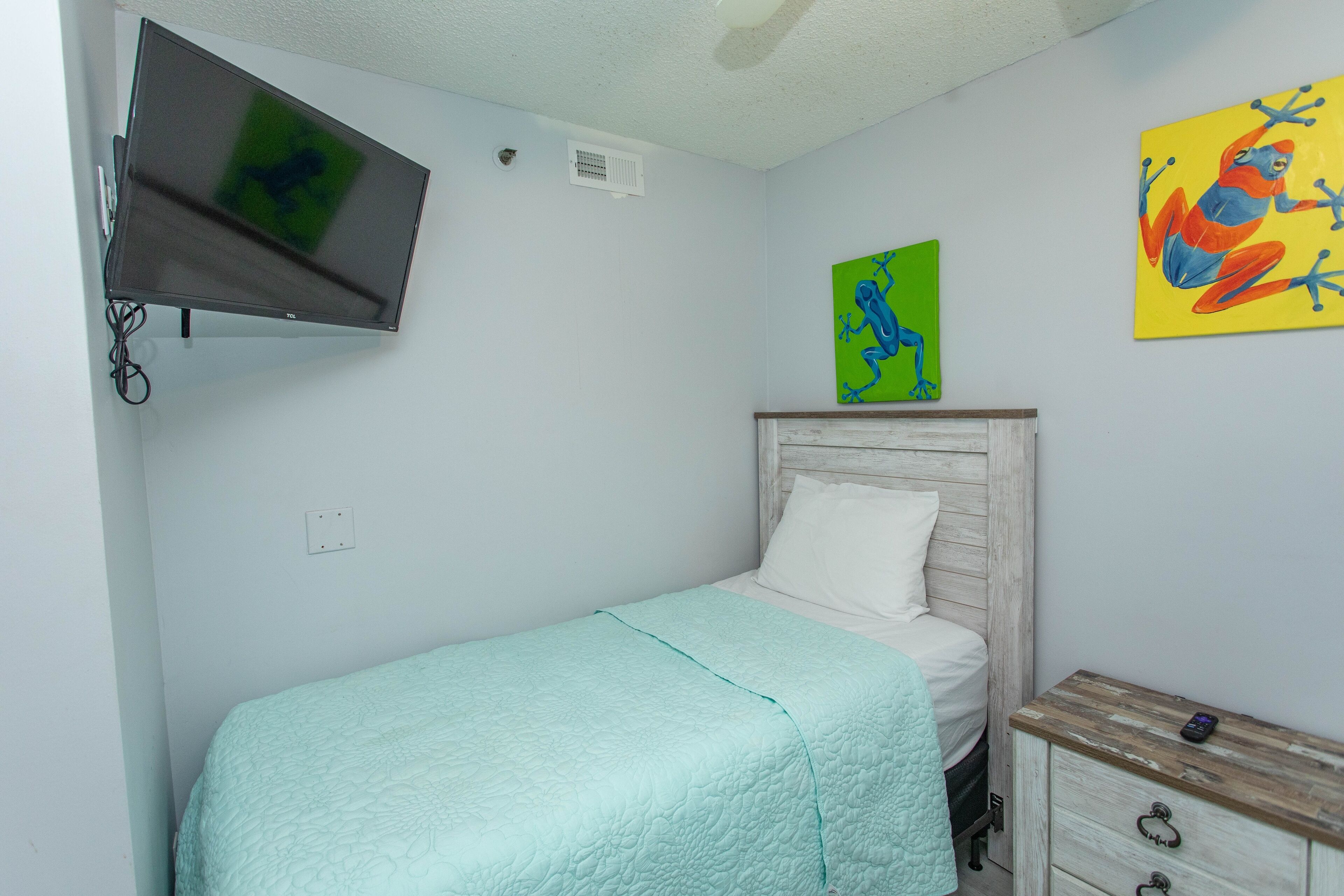 A small, neatly arranged bedroom with a bed, a TV mounted on the wall, and two paintings on the wall.