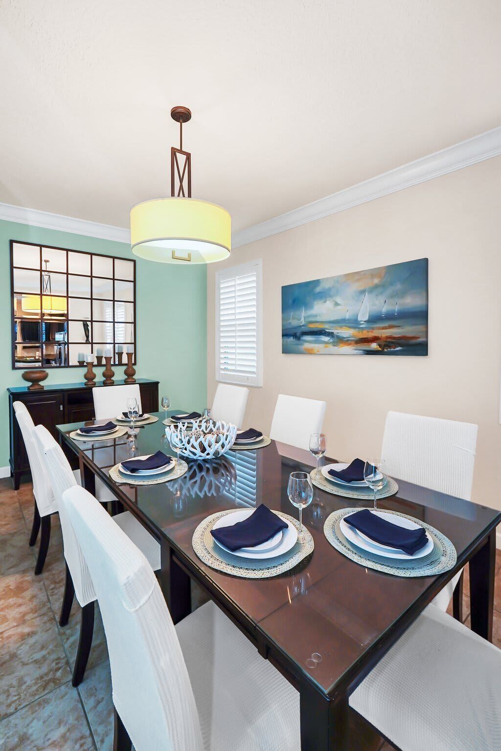 Une salle à manger avec une table en bois foncé dressée pour quatre personnes, des chaises blanches, un lustre, un tableau représentant un paysage marin et une fenêtre avec des stores.