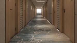 En lång, smal hotellkorridor med ett mönstrat golv, trädörrar och infälld belysning.