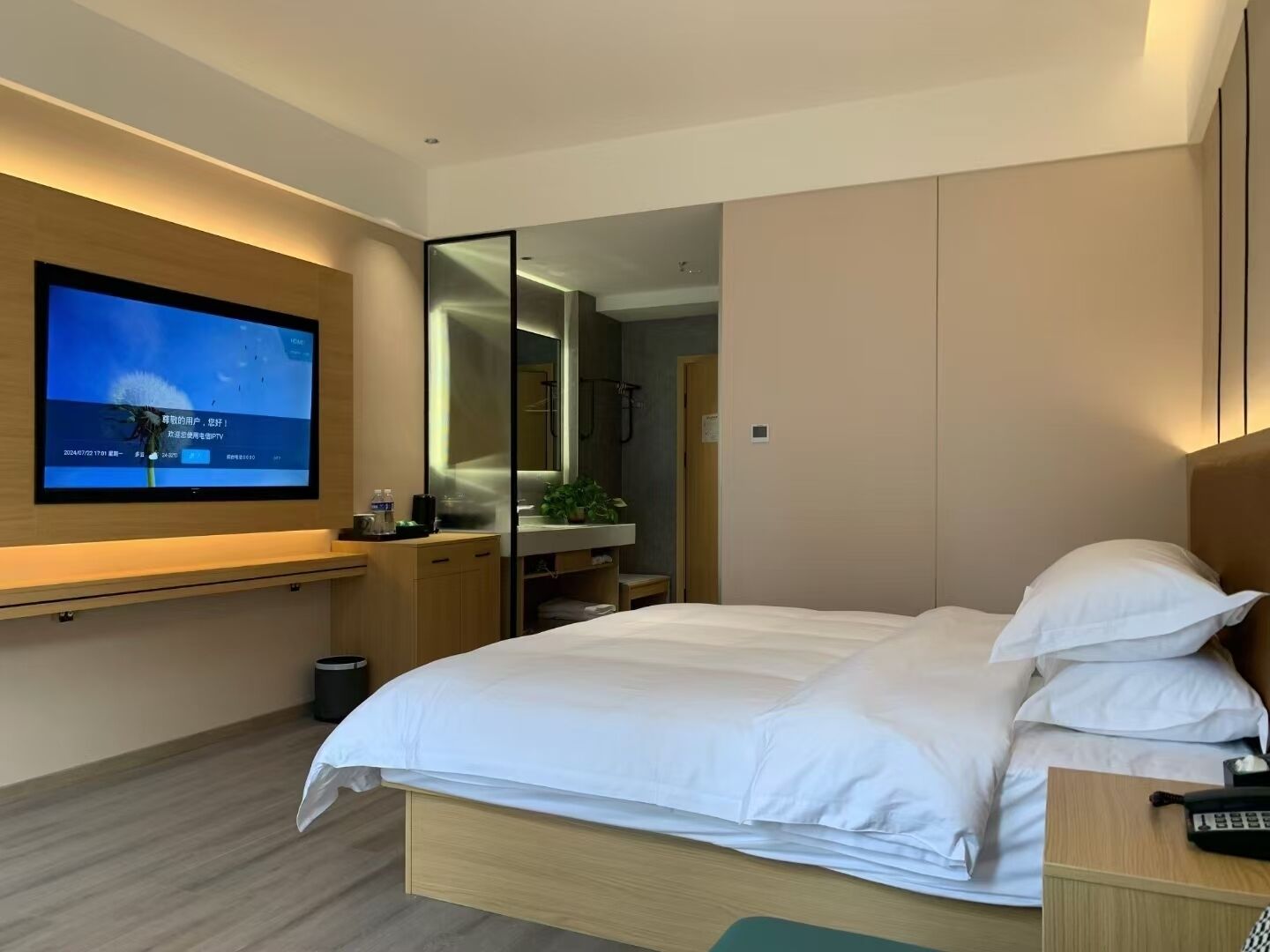 A modern hotel room with a large bed, a TV mounted on the wall, a small desk, and a mirror.
