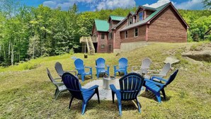 Een houten hutje met een groen dak, omgeven door een gazon met blauwe Adirondack-stoelen rond een vuurkuil.