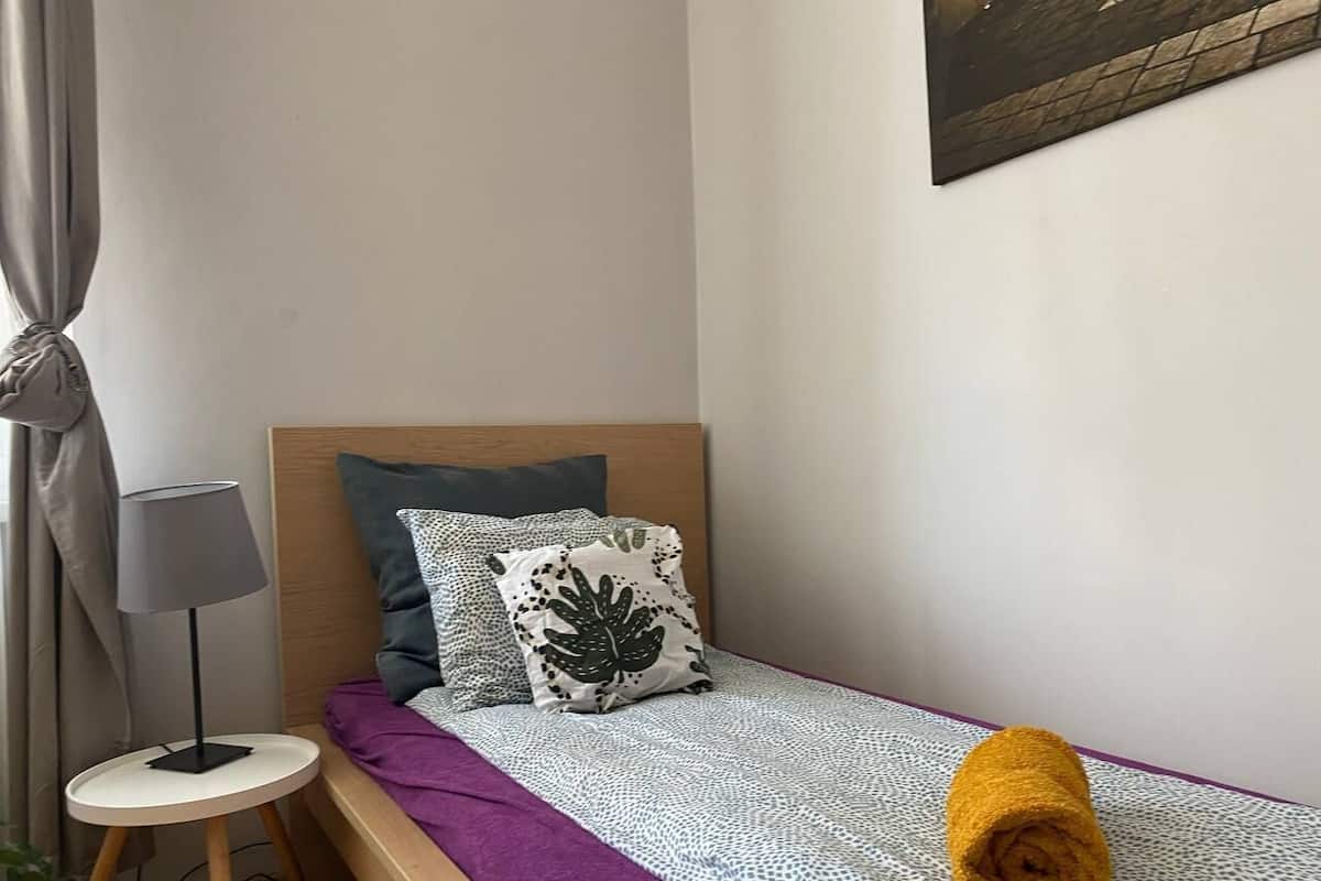Ein kleines, ordentlich bezogenes Bett mit einer violetten Decke, einem gemusterten Überwurf und dekorativen Kissen.