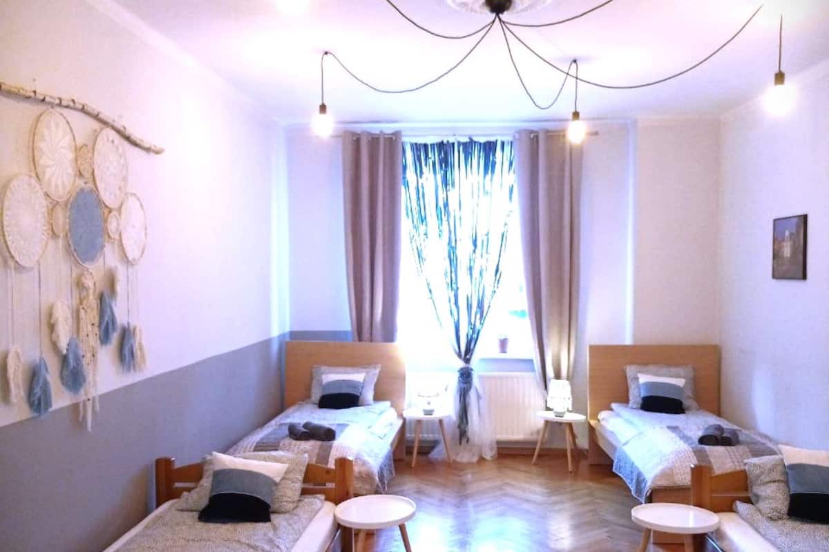 Ein Hotelzimmer mit vier Einzelbetten, die in einer U-Form angeordnet sind, einem Holzboden im Fischgrätmuster, einem großen Fenster mit Vorhängen und einer Wand mit dekorativen Tellern.