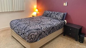 A bedroom with a bed, bedside table, lamp, and a trash bin.