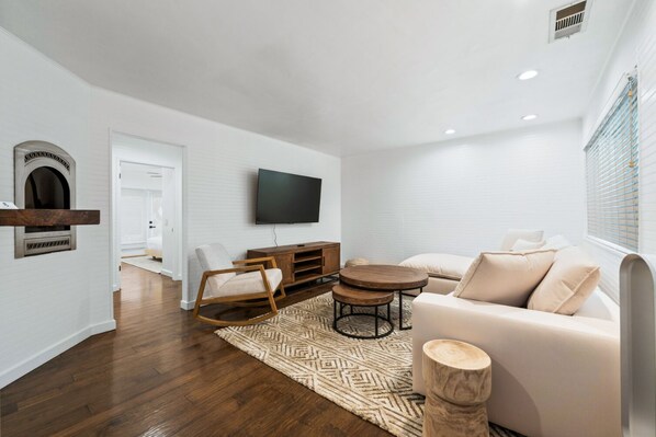 A modern living room with a white sofa, a wooden rocking chair, a coffee table, and a flat-screen TV mounted on the wall.