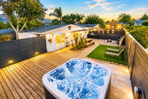 A backyard with a hot tub, wooden deck, and a house with outdoor lighting.