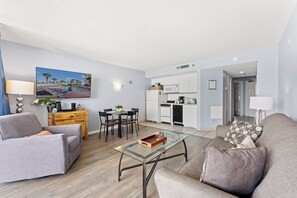 A modern living room with a glass coffee table, grey sofas, a dining area with a table and chairs, a kitchen with white cabinets, and a painting on the wall.