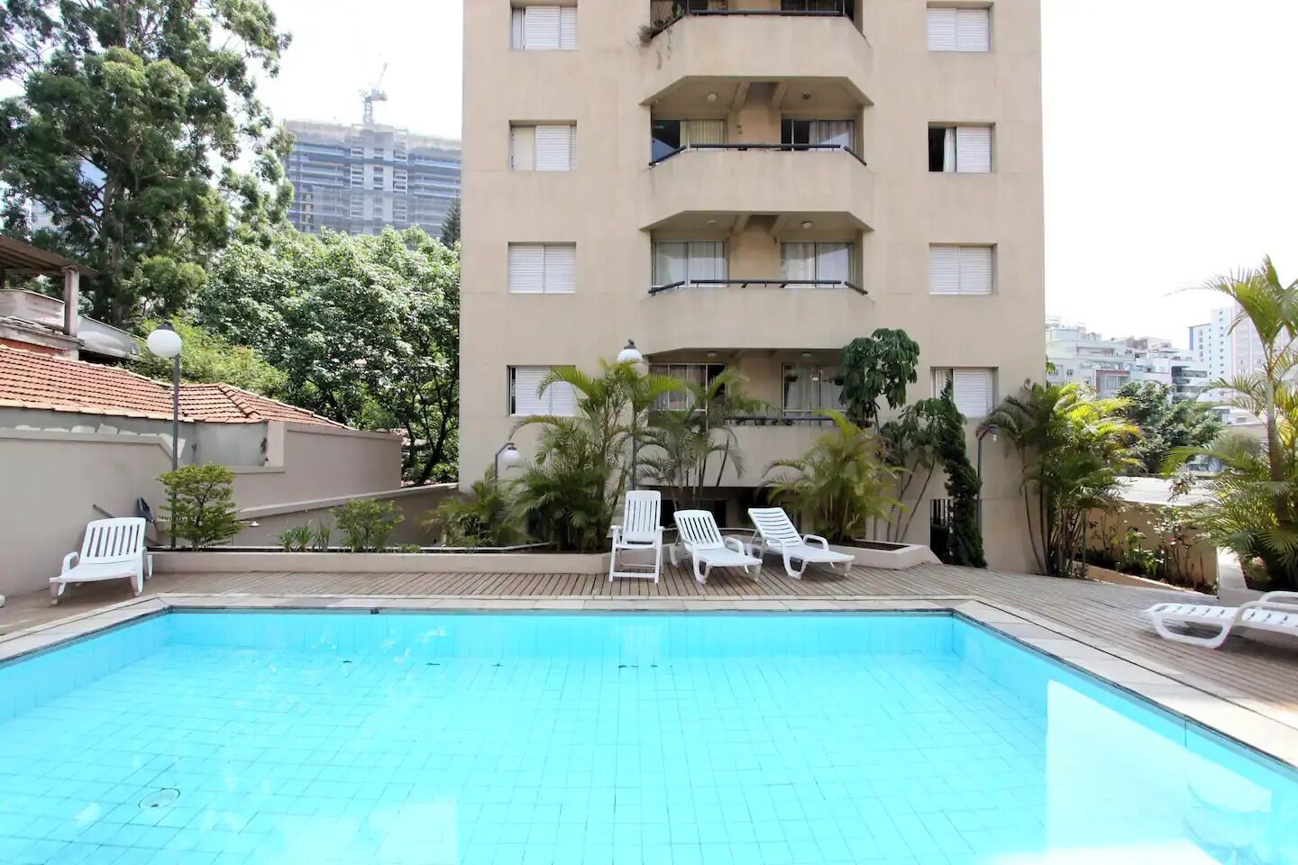 A swimming pool with lounge chairs, surrounded by greenery and a multi-story building in the background.