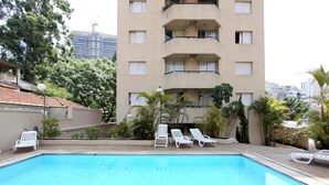 Ein Swimmingpool mit Liegestühlen, umgeben von Grünflächen und einem mehrstöckigen Gebäude im Hintergrund.