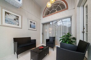 A covered outdoor seating area with wicker furniture, a glass door leading to the interior, and framed pictures on the wall.