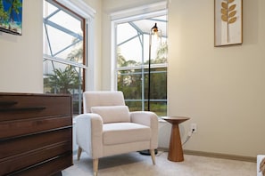 Quarto com uma grande janela, uma poltrona branca, uma cômoda de madeira e uma pequena mesa de madeira.