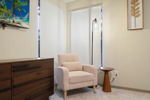 Une pièce comprenant un fauteuil beige, une commode en bois, une petite table ronde et, au mur, une œuvre d’art représentant une feuille, encadrée.