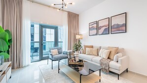 Un salon moderne avec un canapé, un fauteuil et une table basse. Une grande fenêtre avec des rideaux, une plante et une œuvre d’art ornent le mur.