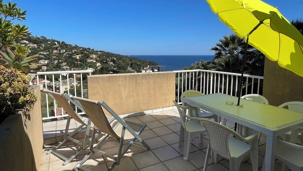 Uma varanda com mesa e cadeiras, um guarda-sol amarelo e vista para o mar e colinas.