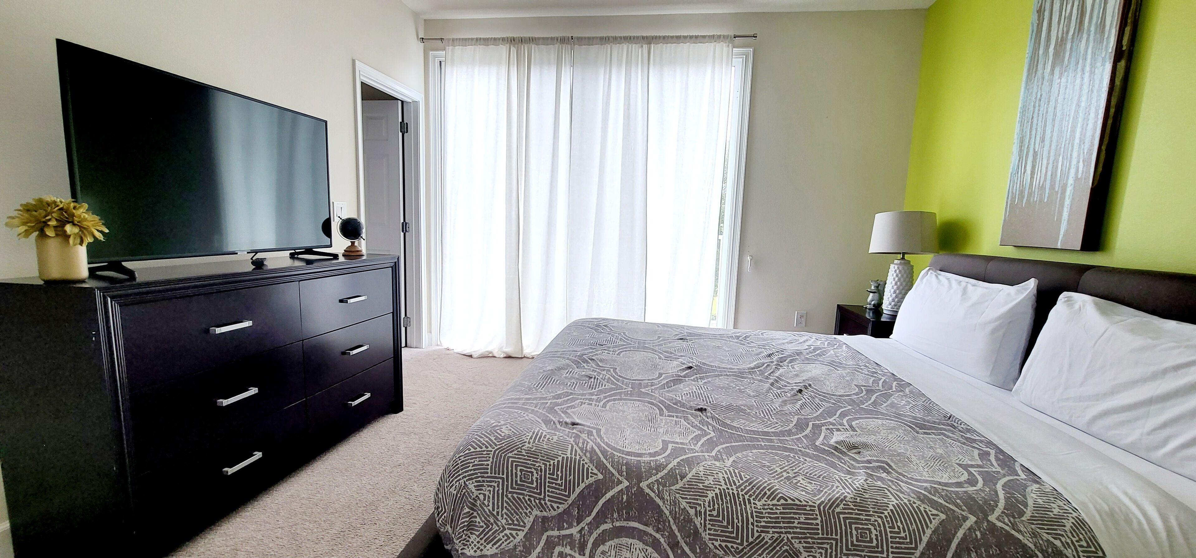 A hotel room with a large bed, a dresser, a flat-screen TV, and a window with curtains.