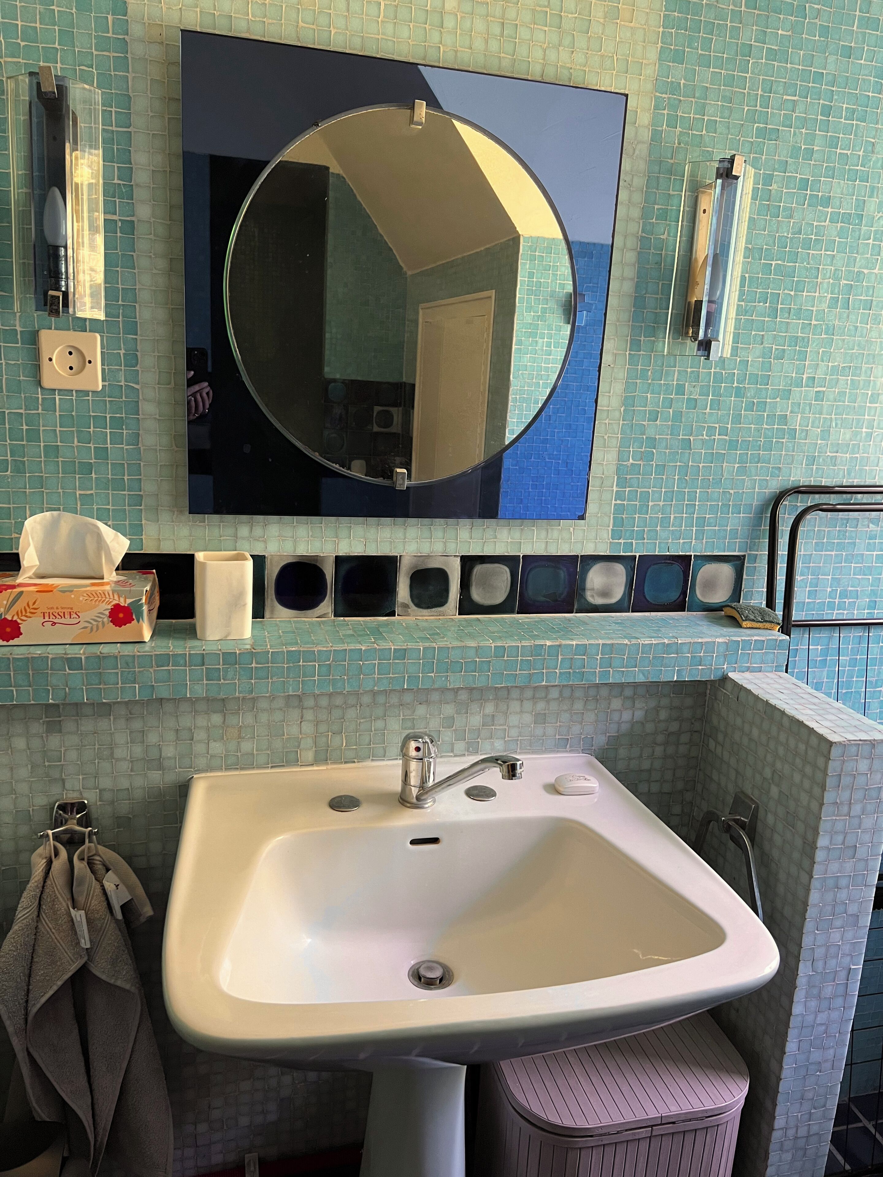 Ein Badezimmer mit einem weißen Waschbecken, einem runden Spiegel und einer gefliesten Rückwand.