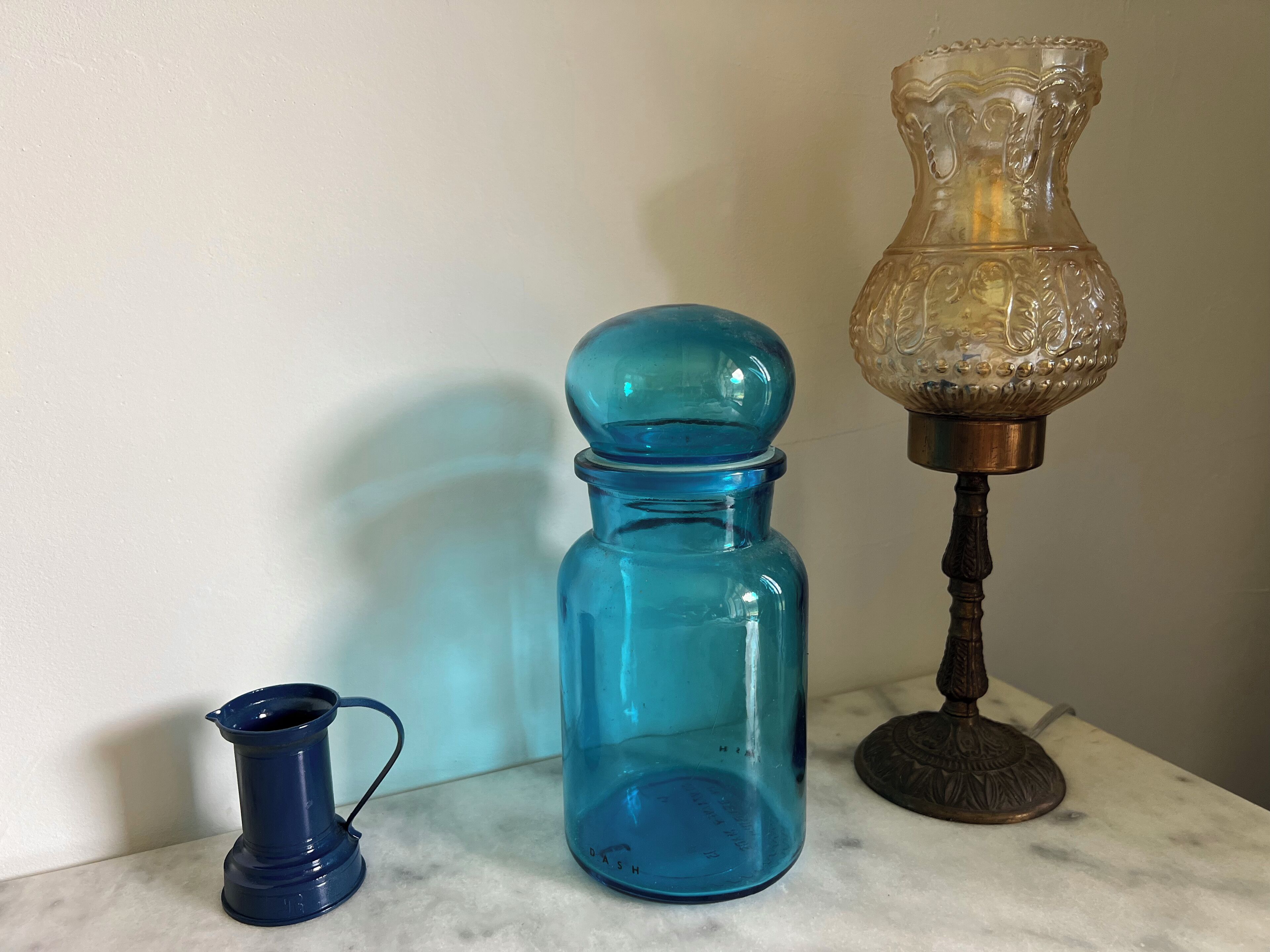 A blue glass jar, a smaller blue glass pitcher, and a decorative glass lamp on a marble surface.