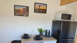 A compact kitchen with a microwave, refrigerator, two bar stools, a small table with a plant, and two framed pictures on the wall. - Gem View - Tiny home (Barwite)