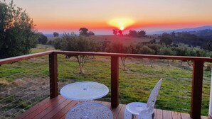 A wooden deck with two white metal chairs and a table, overlooking a grassy landscape with trees and a sunset. - Gem View - Tiny home (Barwite)