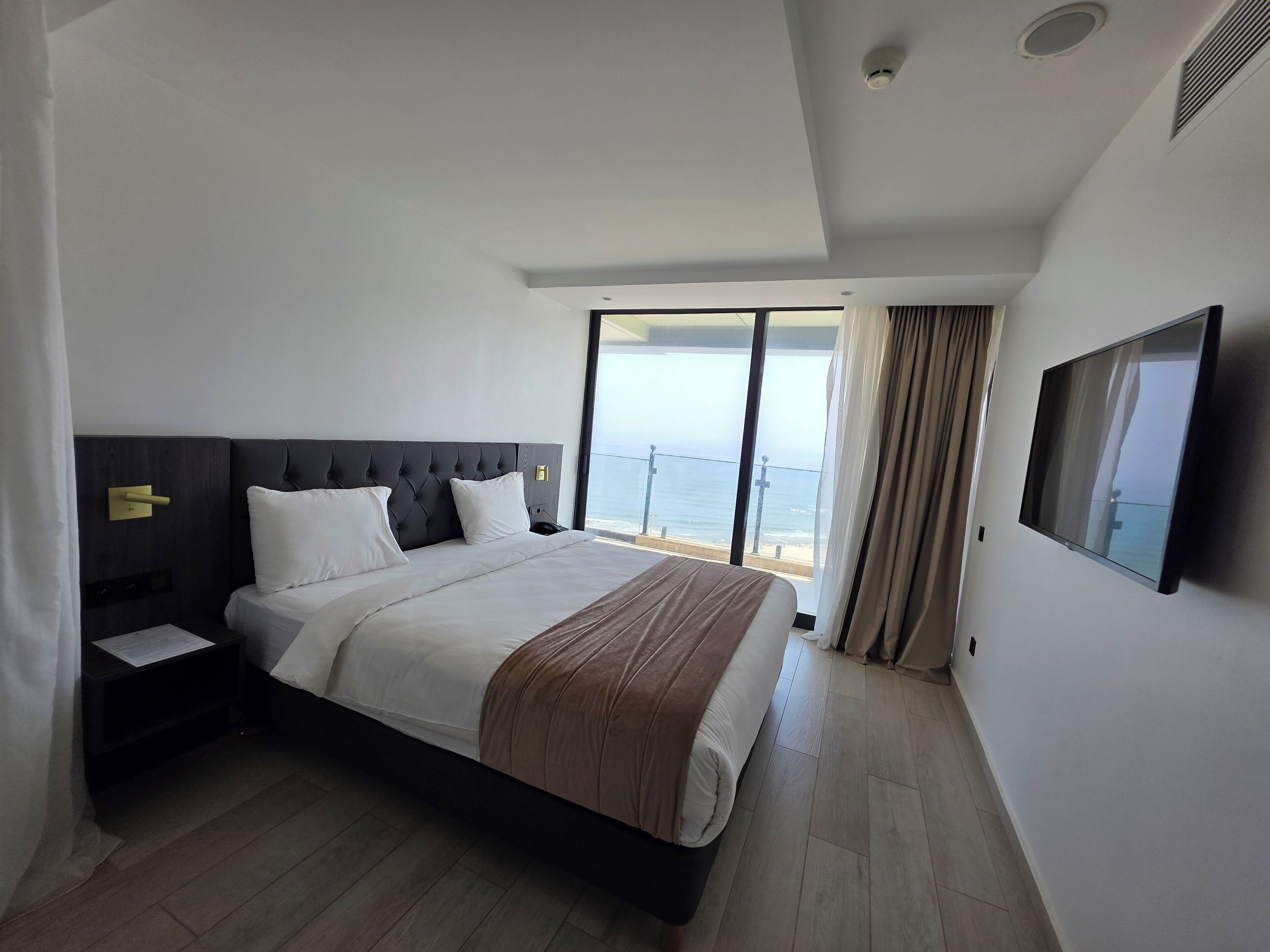 A modern hotel room with a large bed, a flat-screen TV, and a balcony with a view of the sea.