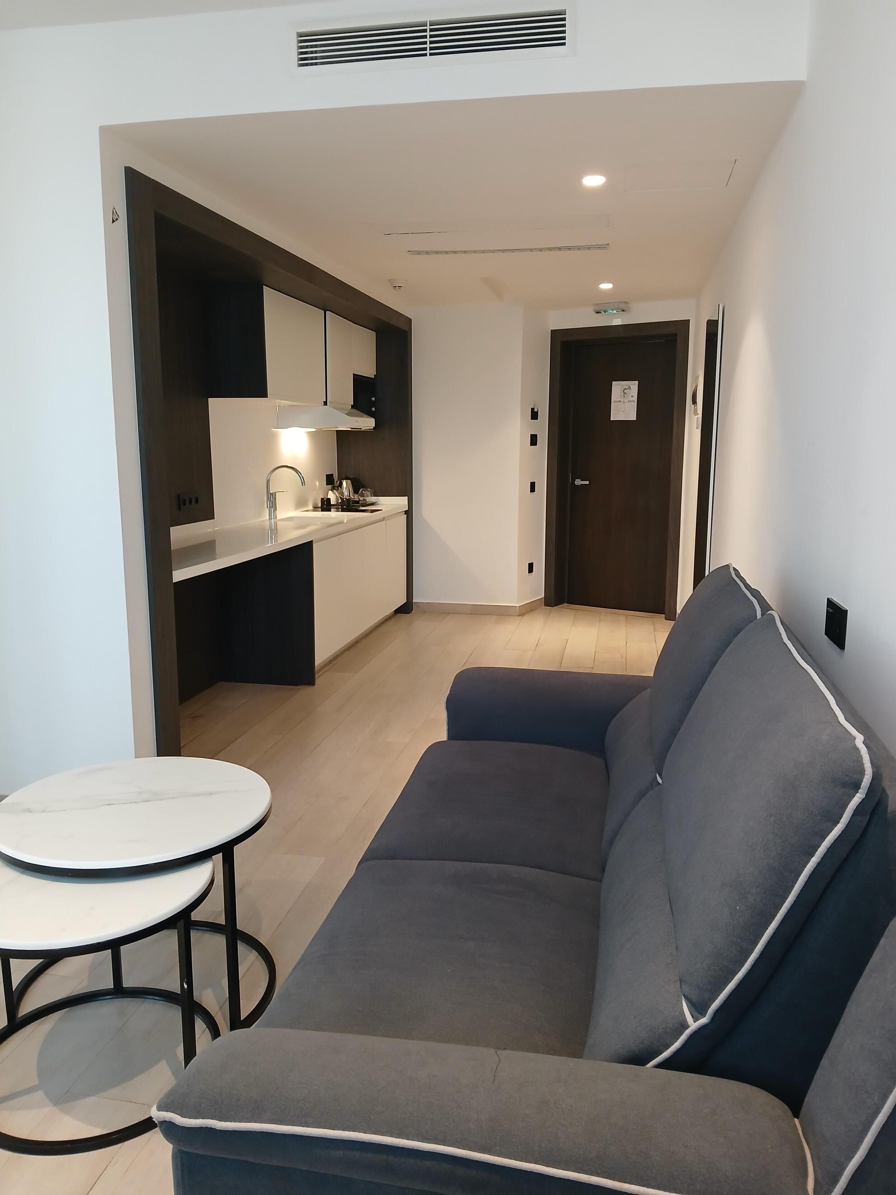 A modern living room with a grey sofa, a small round table, and a kitchenette area with a white countertop and dark cabinets.