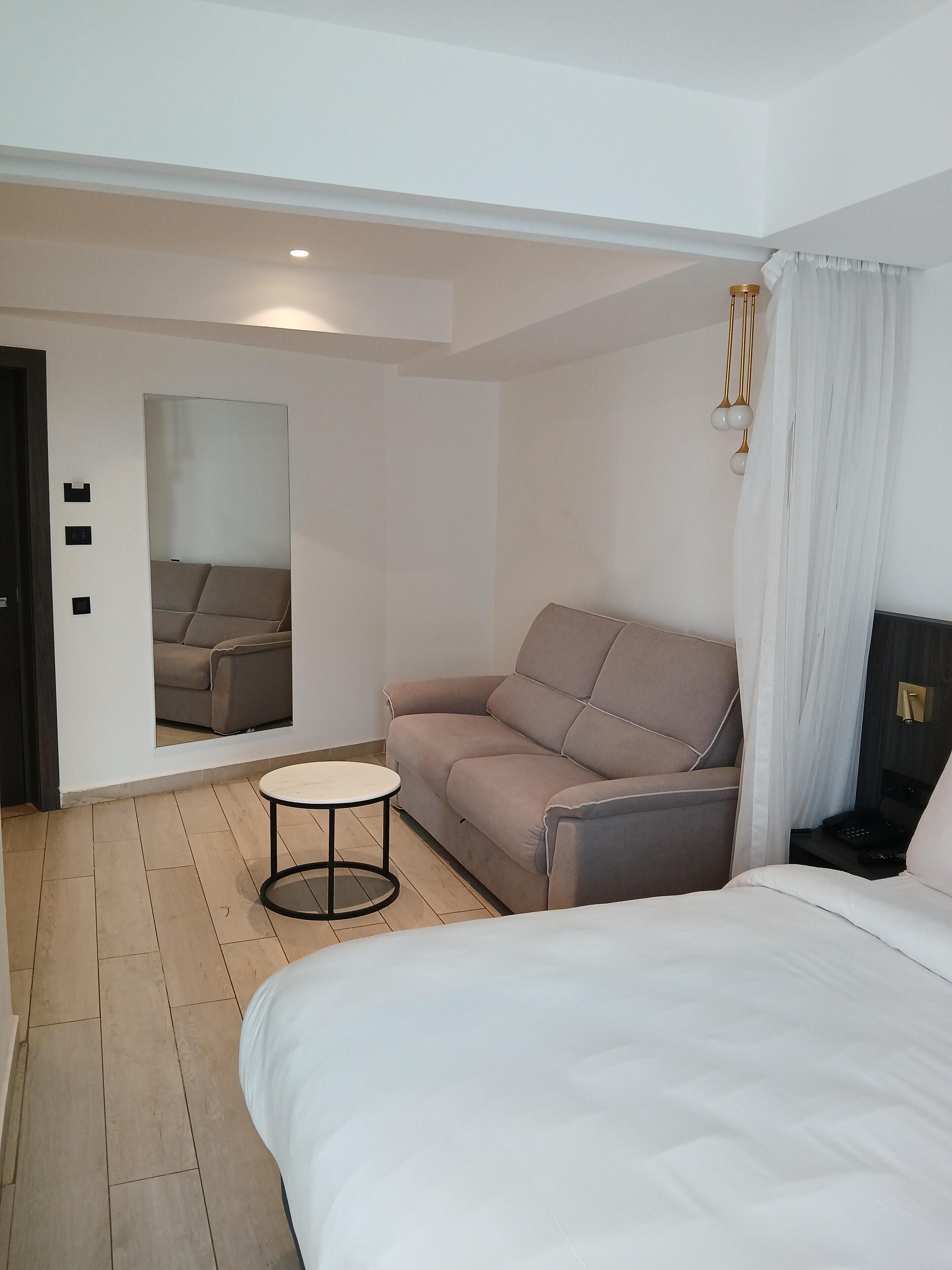 Habitación de hotel moderna con una cama grande, un sofá, una mesita redonda y un espejo que refleja la habitación.