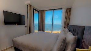 A hotel room with a large bed, a flat-screen TV, and a view of the sea through large windows.
