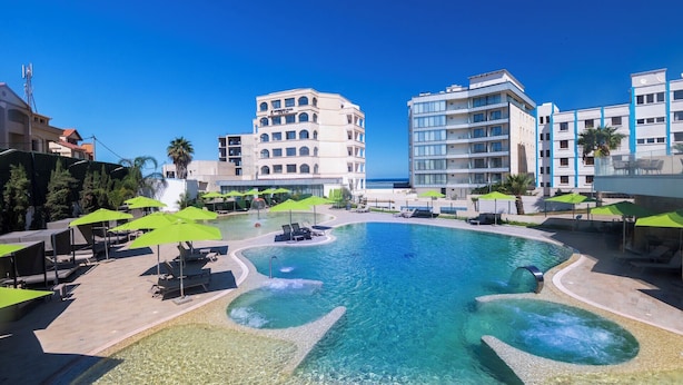 A resort pool with clear water, surrounded by lounge chairs and umbrellas, and adjacent to modern buildings.