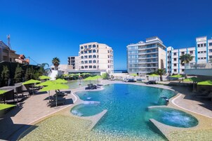 A resort pool with clear water, surrounded by lounge chairs and umbrellas, and adjacent to modern buildings.