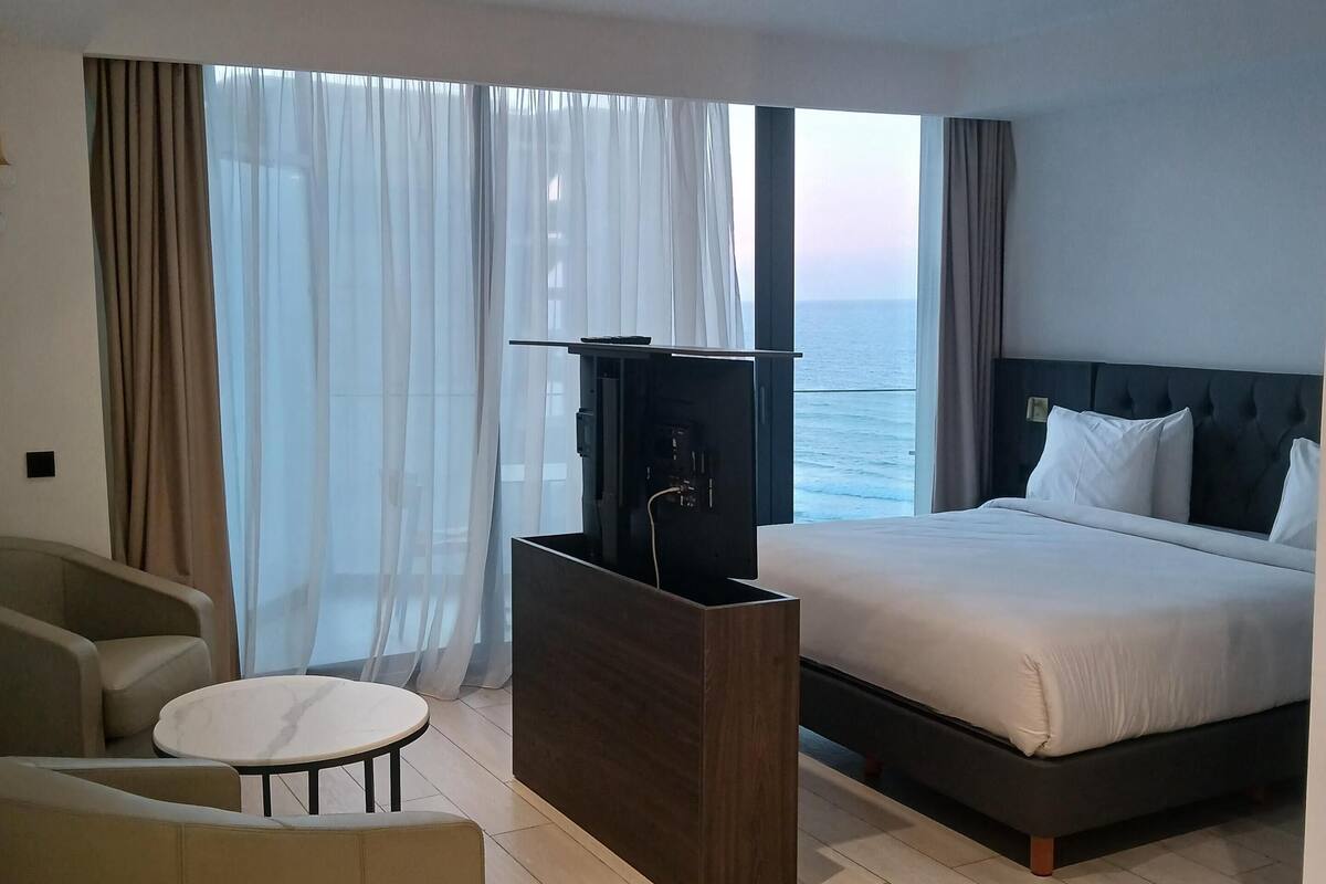 A modern hotel room with a large bed, a seating area with a round table, and a view of the sea through large windows.