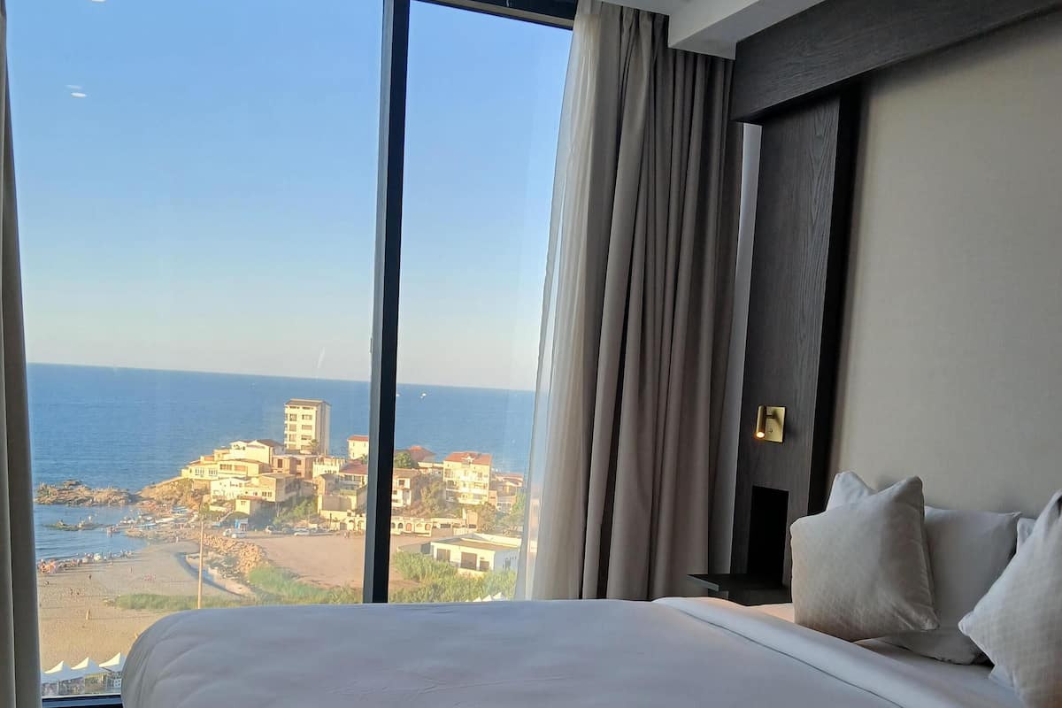 A hotel room with a large bed, a nightstand, and a view of the sea and buildings.