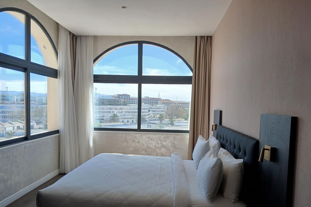 A hotel room with a large bed, a headboard, and a view of buildings through a large window.