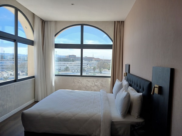 A hotel room with a large bed, a headboard, and a view of buildings through a large window. - Hôtel Boumerdes Plaza (Boumerdès)