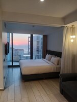 A modern hotel room with a large bed, a sofa, a TV, and a balcony with a view of the sunset and buildings.