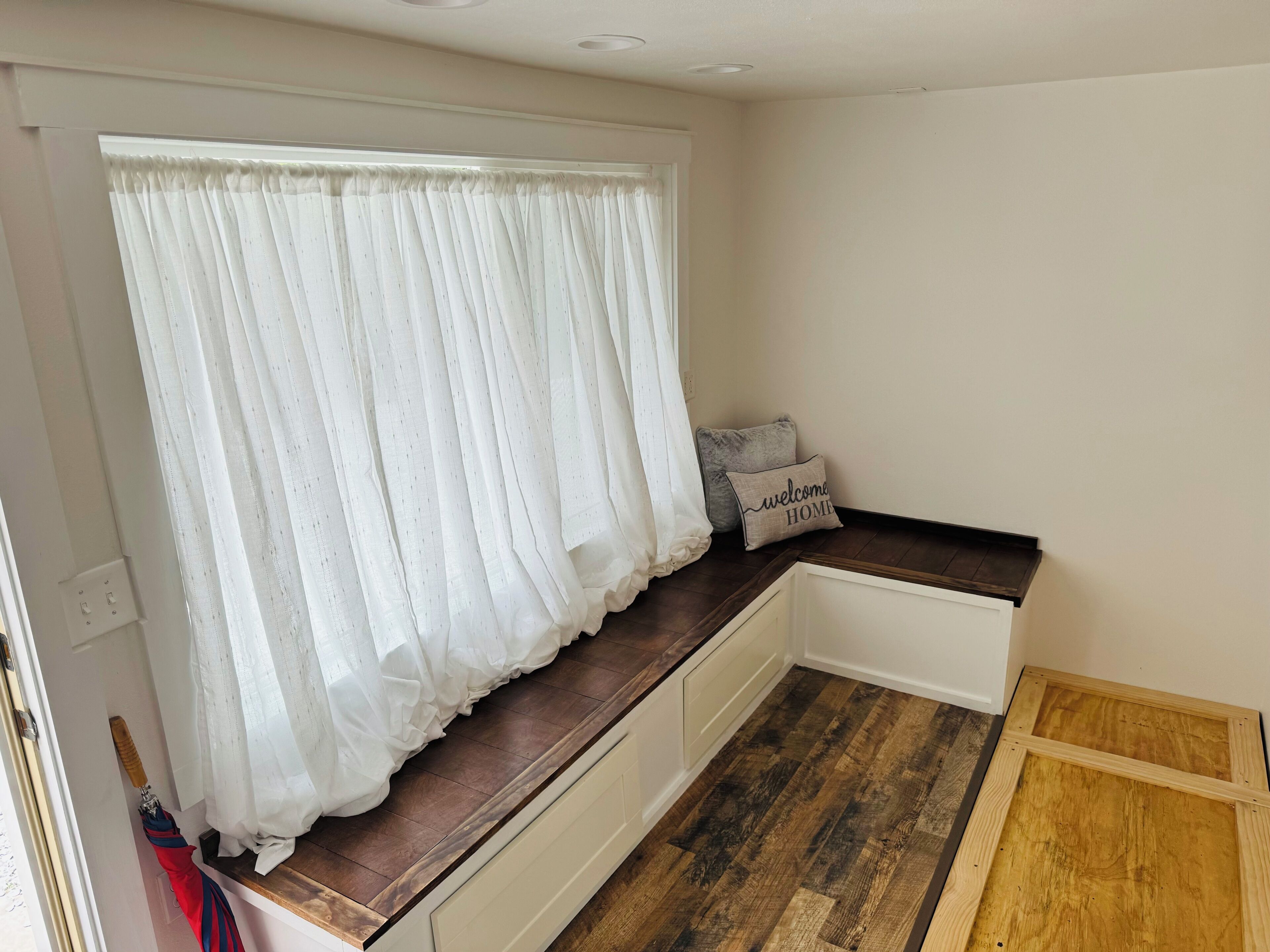 A room with a window covered by sheer white curtains, a small bench with a cushion, and a wooden floor.