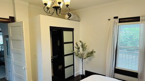 Een slaapkamer met een groot bed, een kroonluchter, een deur, een raam met jaloezieën en een kleine plant.