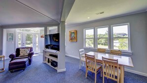 Een ruime woonkamer met een grote televisie, een eethoek met houten stoelen en grote ramen met uitzicht op de omgeving.