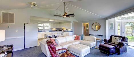 A spacious living room with a white sectional sofa, a red leather armchair, and a dark leather armchair. A ceiling fan and a large window provide natural light.