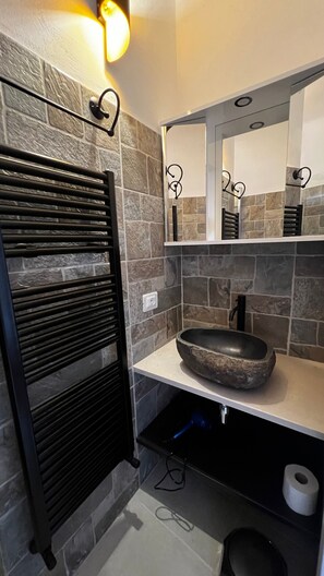 A bathroom with a stone wall, a black towel rack, a sink with a stone basin, a mirror, and a toilet. - Locazione turistica Il Boschetto (Martina Franca)