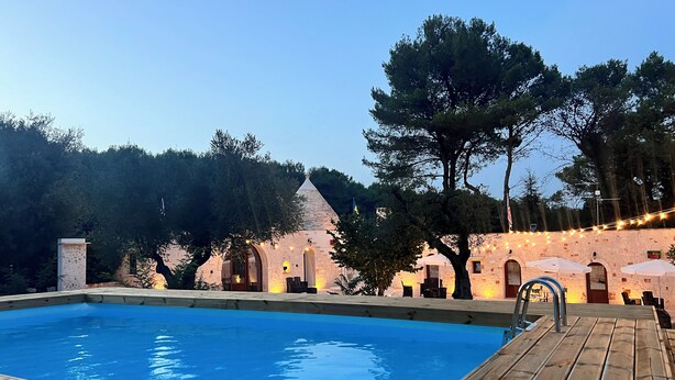 Ein Schwimmbad mit Holzterrasse, ein Steinhaus mit Rundbogenfenstern und Lichterketten.