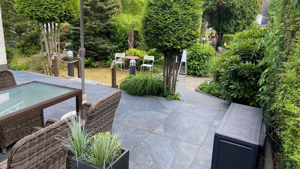Une terrasse extérieure avec des meubles en osier, une table en verre et un pot noir contenant des plantes vertes.