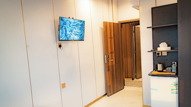 Una habitación con una cama, un televisor montado en la pared, un armario de madera y una pequeña kitchenette con refrigerador.