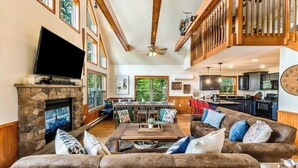 A spacious living room with a stone fireplace, a flat-screen TV, a dining area with a high ceiling, and a kitchen in the background.