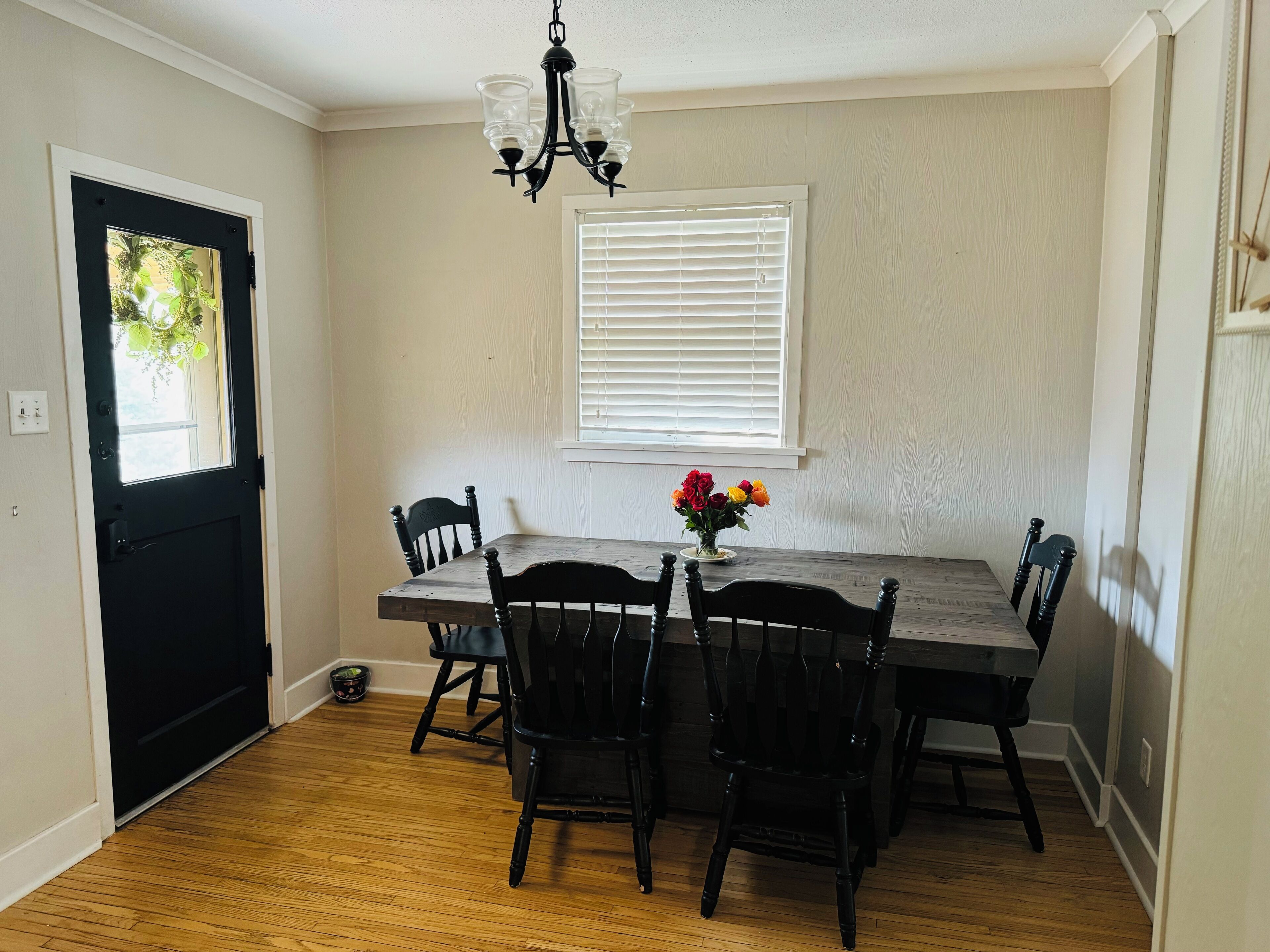 A dining area with a wooden table and chairs, a chandelier, a window with blinds, and a door with a view of greenery.